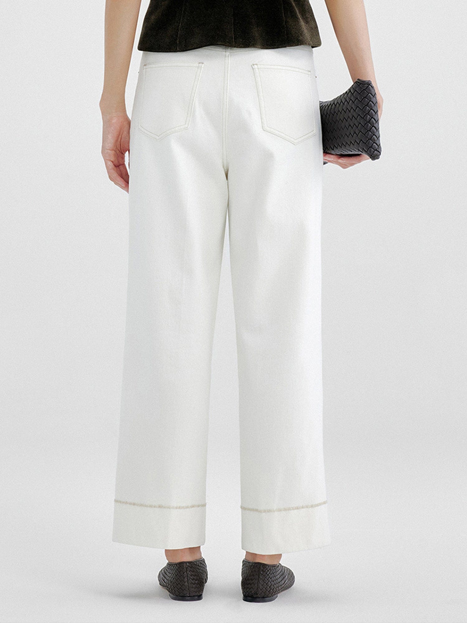 Woman wearing Arkin Studio tailored cropped jeans with topstitching in white and olive green top, wearing a pair of black shoes, holding a back bag, standing facing a white wall, on a white background