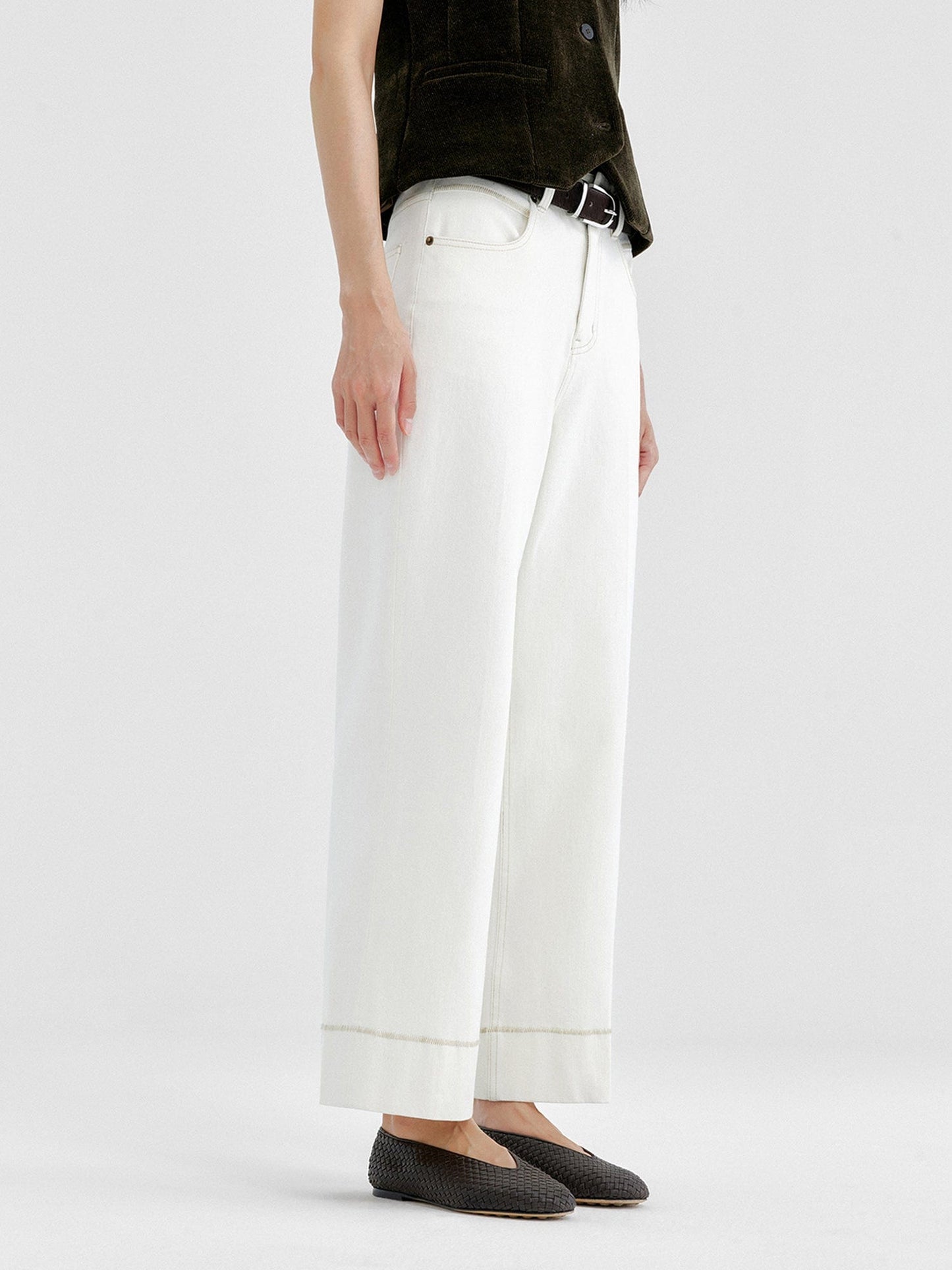 Woman wearing Arkin Studio tailored cropped jeans with topstitching in white and olive green top, wearing a pair of black shoes, standing sideways, on a white background
