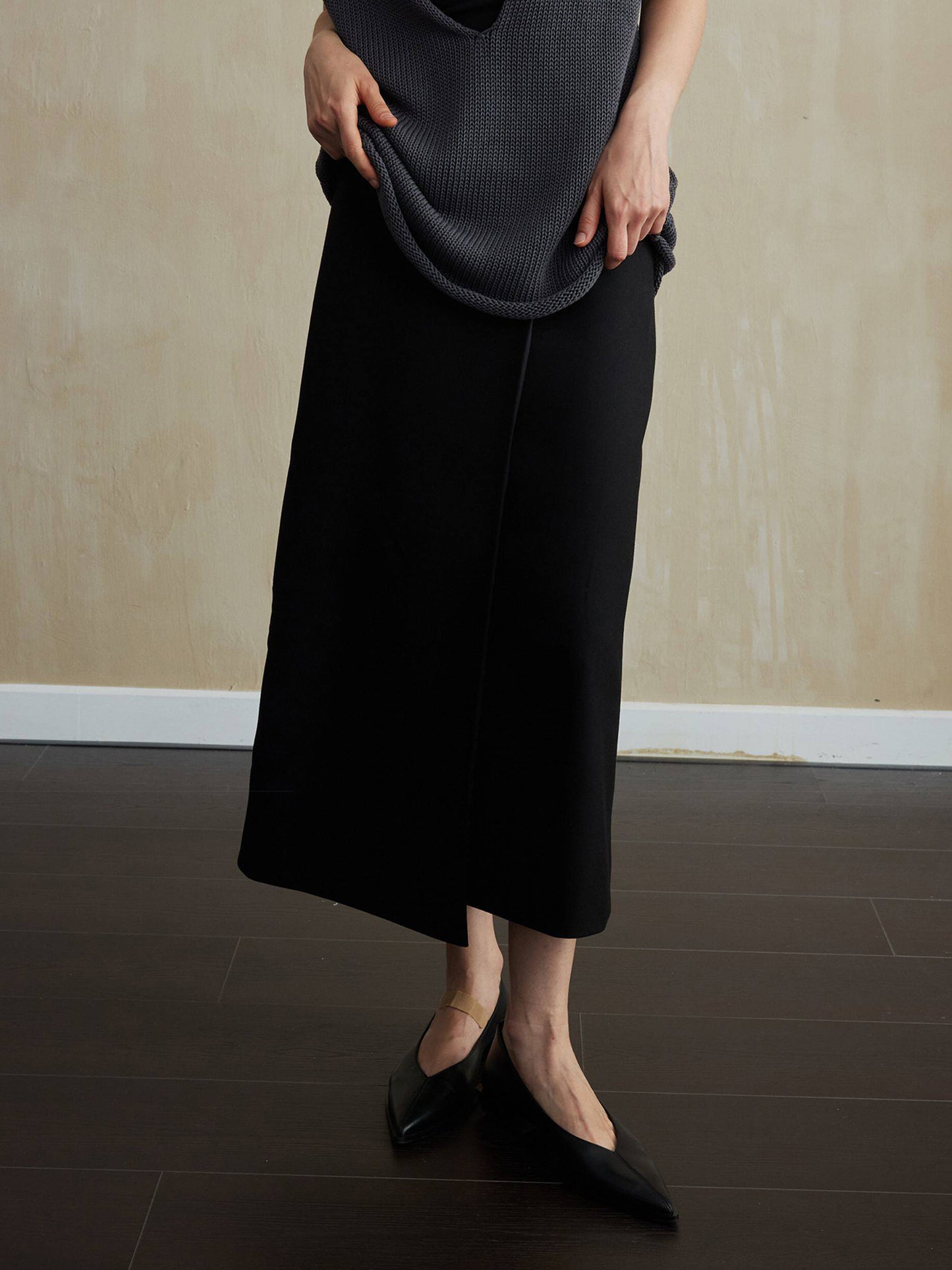 Woman wearing Arkin Studio tencel wool twill a-line skirt in black and a dark gray sleeveless knit top, with pointed leather shoes on feet, facing the camera, standing on the dark-brown floor, a yellow wall behind her
