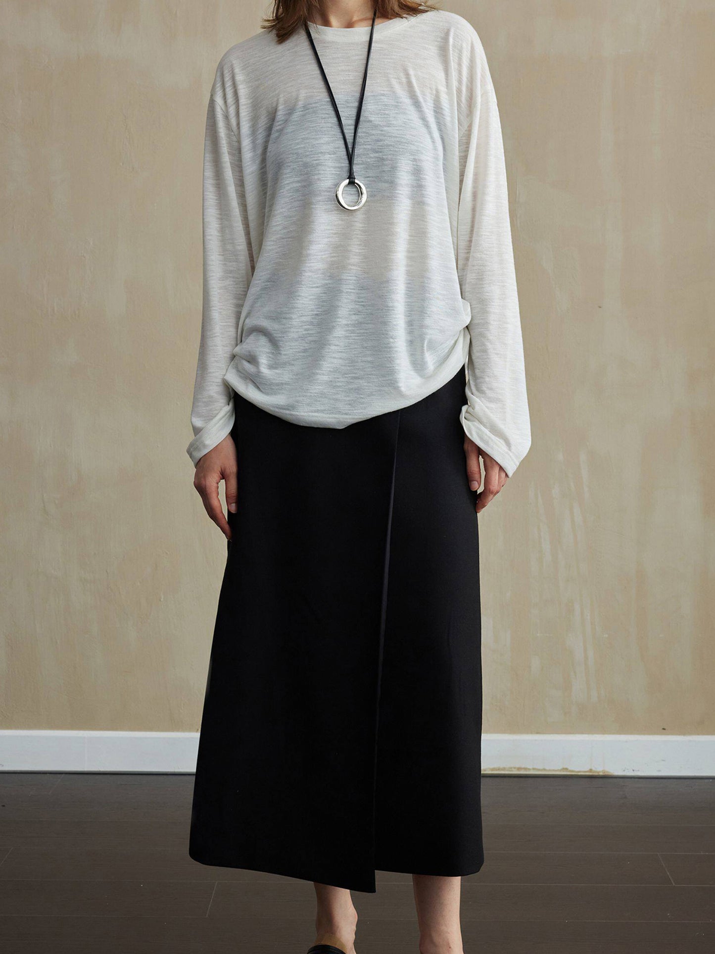 Woman wearing Arkin Studio tencel wool twill a-line skirt in black and a light gray long-sleeve top, facing the camera, standing on the dark-brown floor, a yellow wall behind her