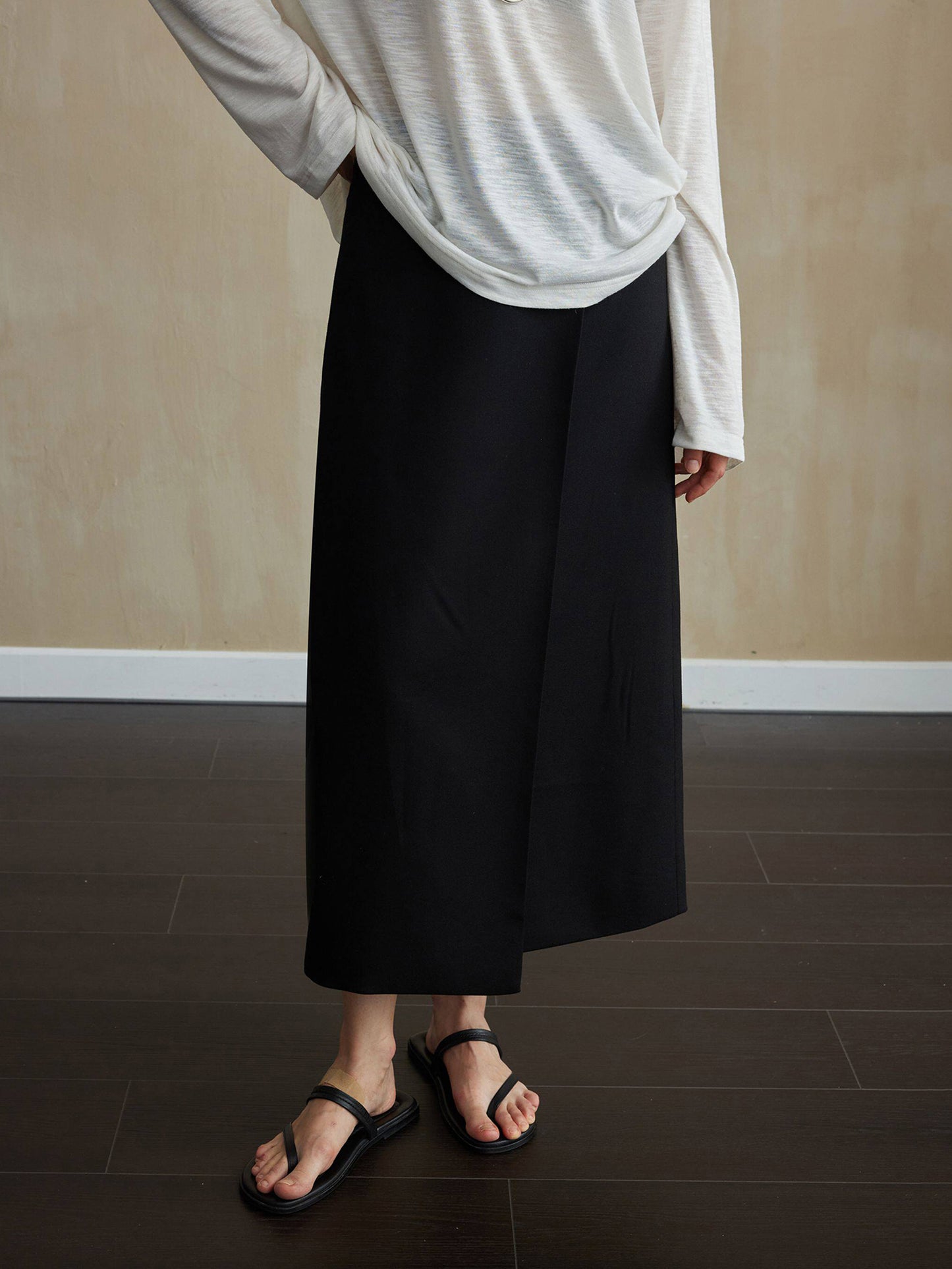 Woman wearing Arkin Studio tencel wool twill a-line skirt in black and a light gray long-sleeve top, with summer slippers on feet, facing the camera, standing on the dark-brown floor, a yellow wall behind her