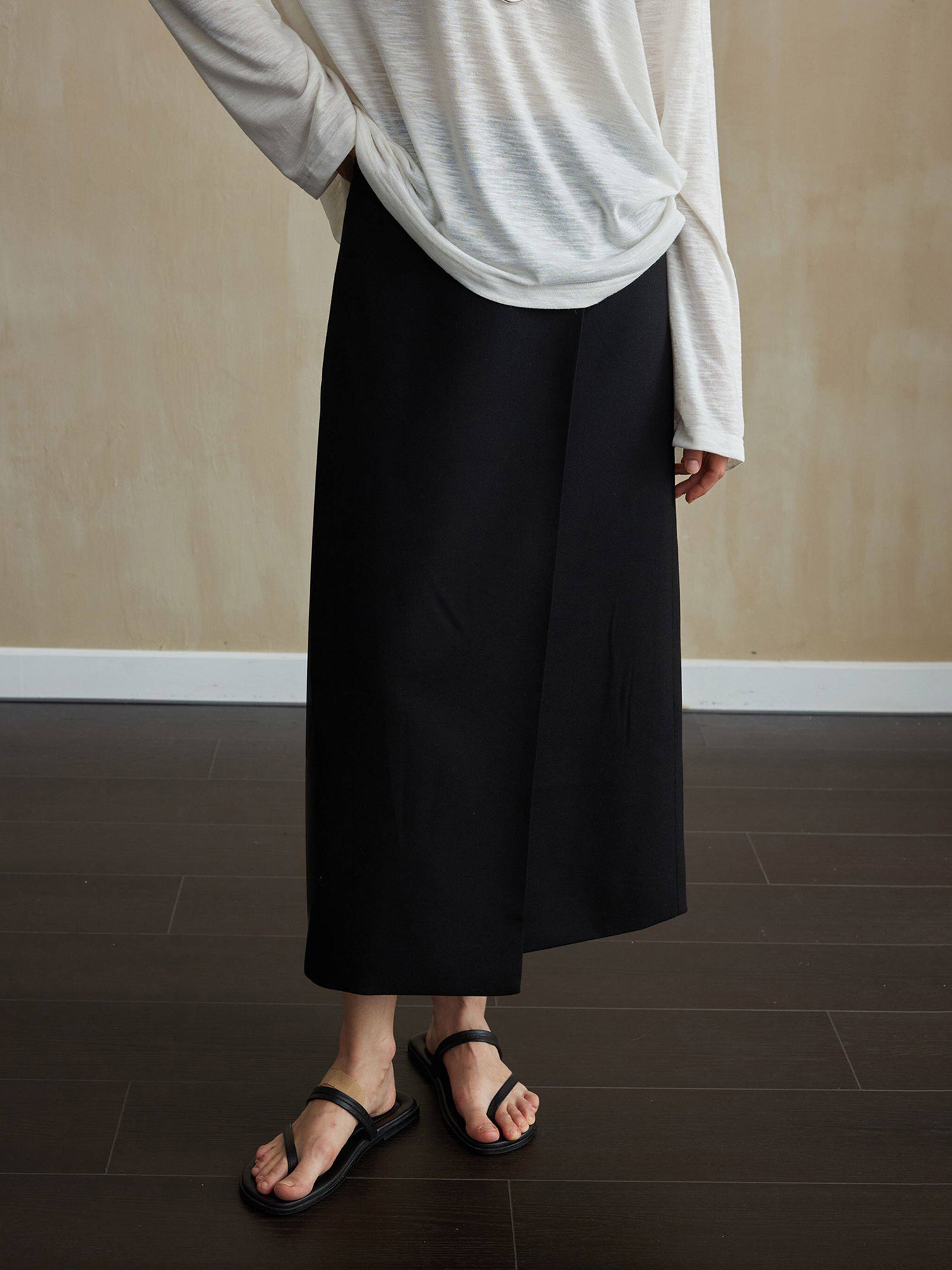 Woman wearing Arkin Studio tencel wool twill a-line skirt in black and a light gray long-sleeve top, with summer slippers on feet, facing the camera, standing on the dark-brown floor, a yellow wall behind her