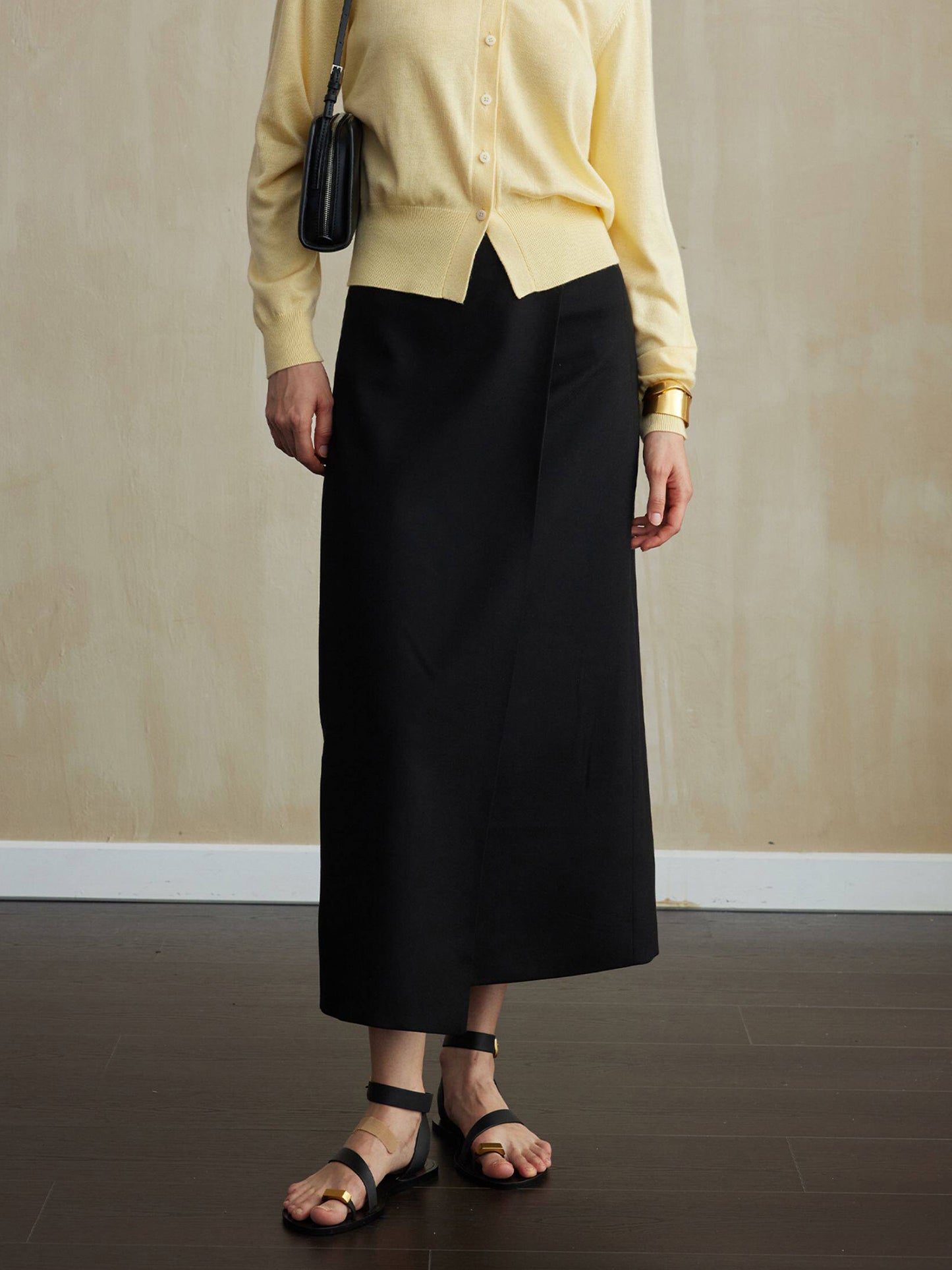 Woman wearing Arkin Studio tencel wool twill a-line skirt in black and a yellow cardigan, with summer sandals on feet, facing the camera on the dark-brown floor, a yellow wall behind her