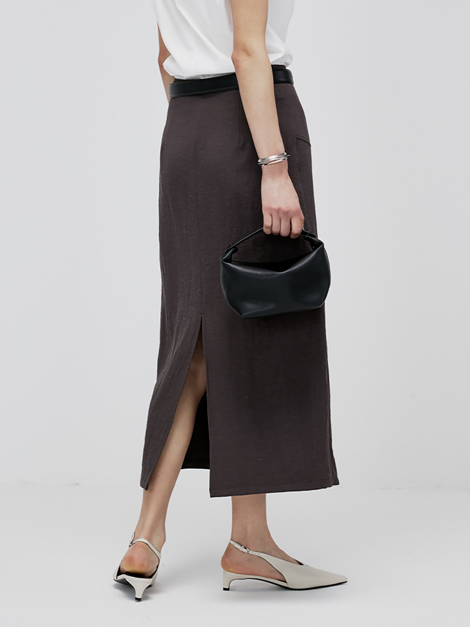 Woman wearing Arkin Studio textured h-line slit midi skirt with resin button in chocolate and a white top, with a black bag in hand, standing with her back to the camera, on a white background