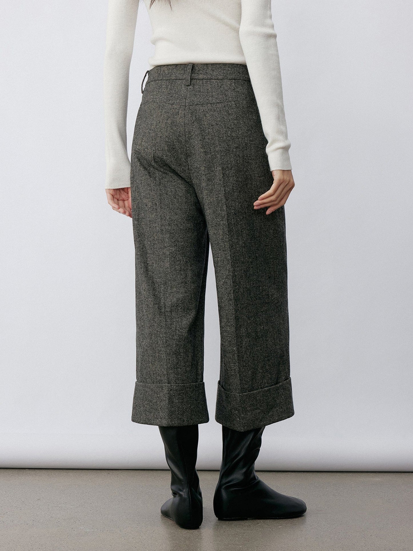Woman wearing Arkin Studio turn-up hem wool trousers in gray and a white long sleeve top, with hands down at her sides, wearing a pair of black shoes, standing facing a white wall on a khaki floor
