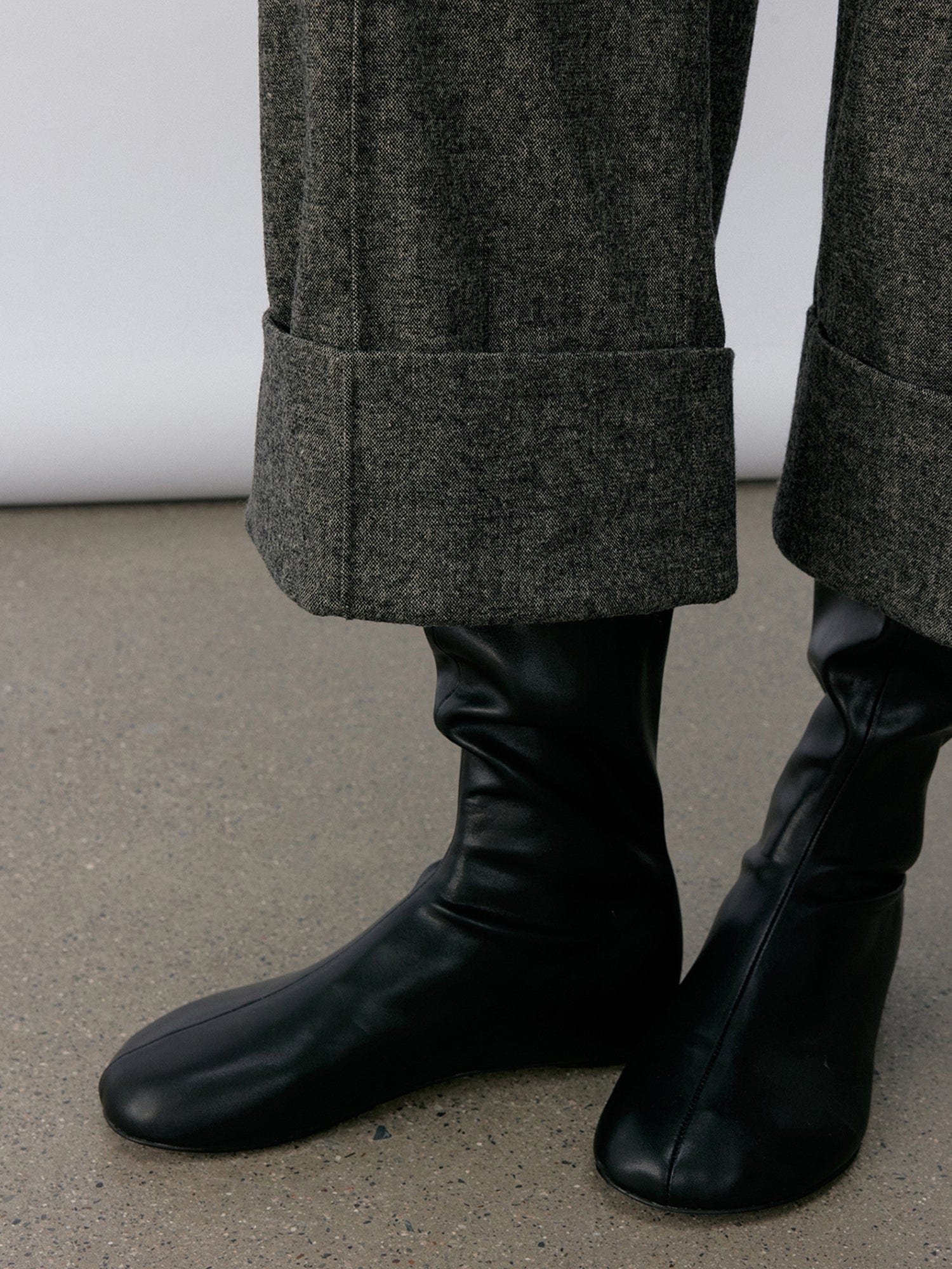 Close-up of a woman's legs from calf to feet wearing Arkin Studio turn-up hem wool trousers in gray with a pair of black shoes, standing sideways, on a khaki floor, in front of a white wall