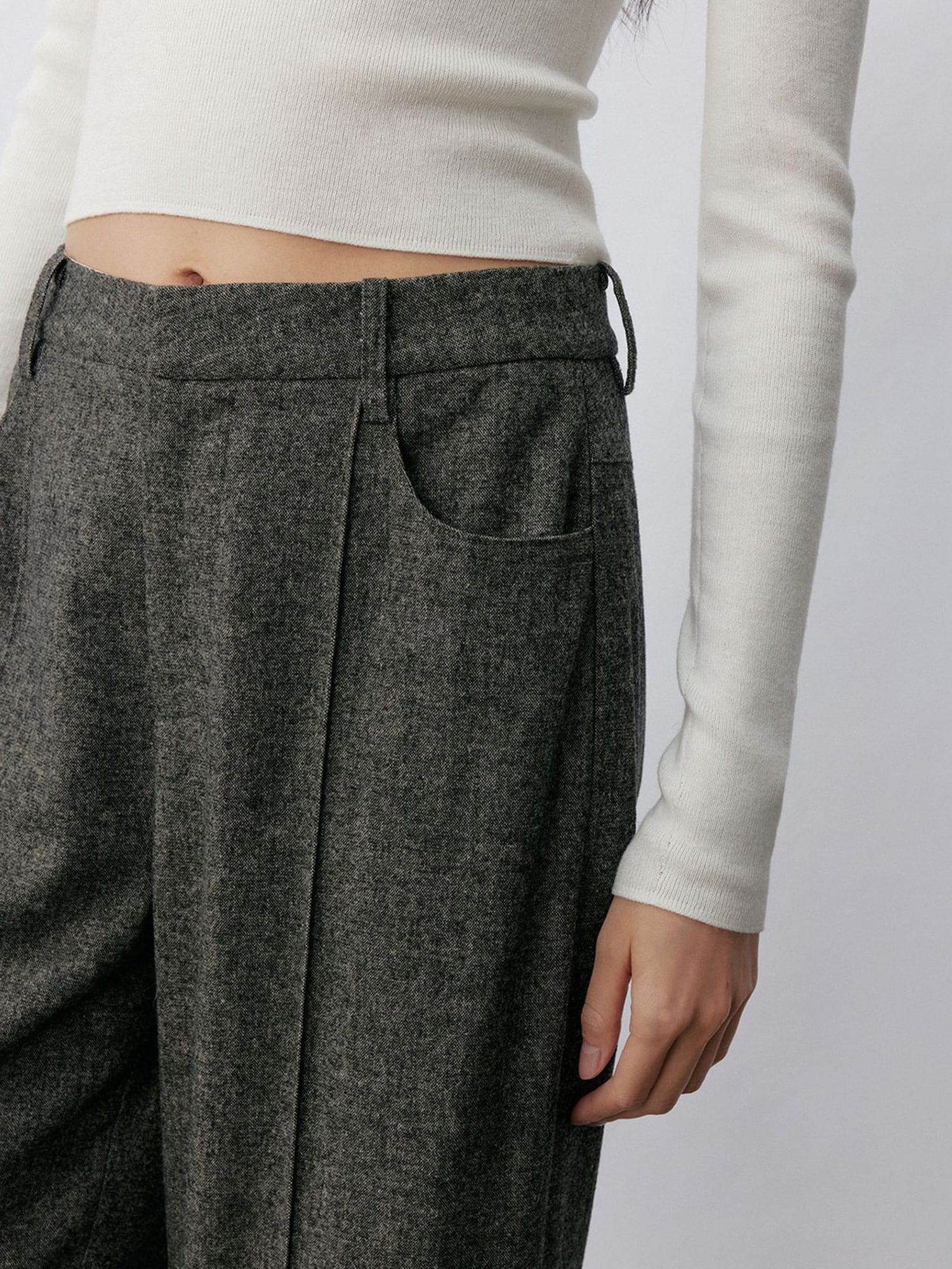 Woman wearing Arkin Studio turn-up hem wool trousers in gray and a white long sleeve top, with one hand down at her side, focus on her waist, standing facing the camera, on a white background