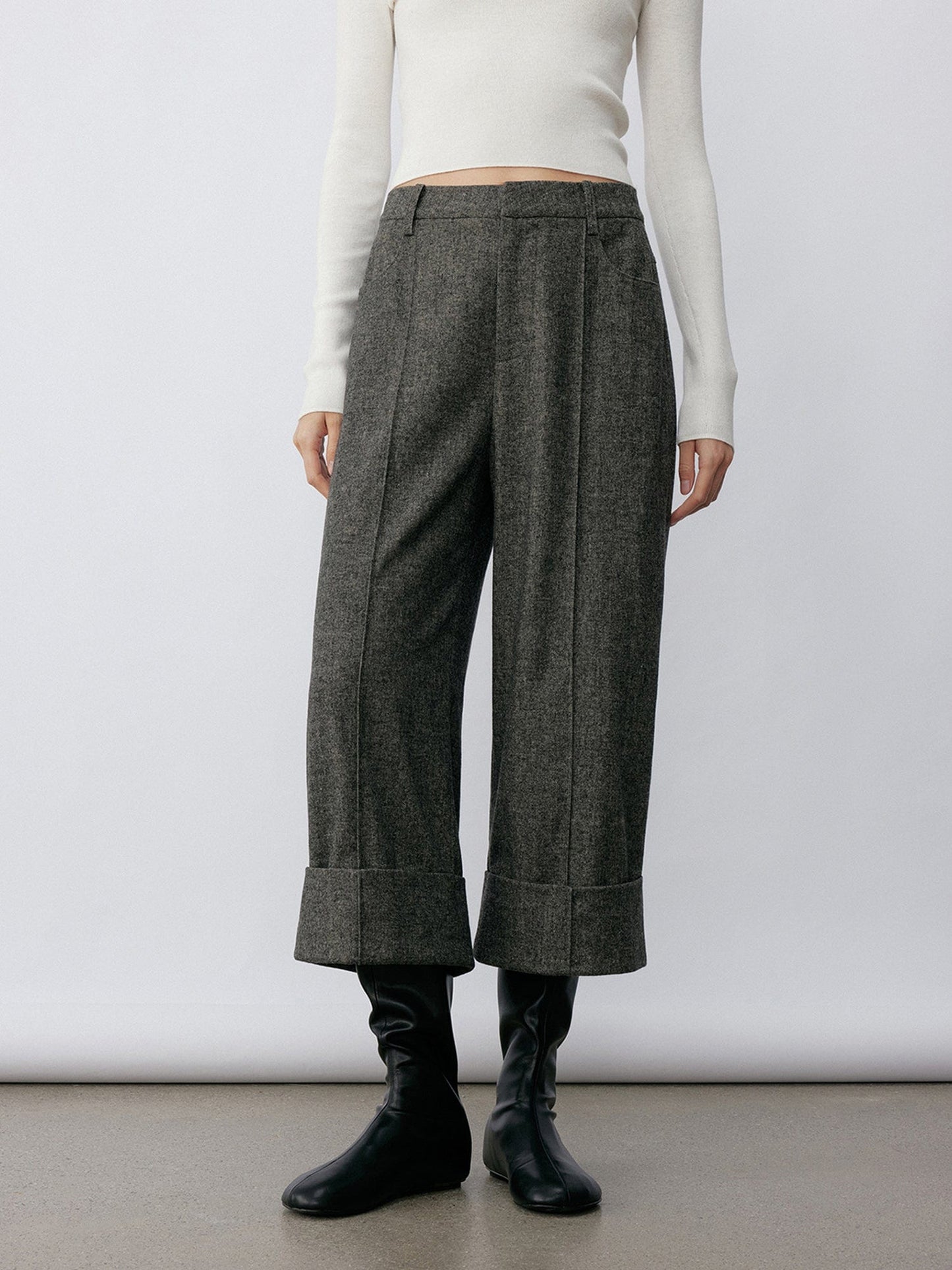 Woman wearing Arkin Studio turn-up hem wool trousers in gray and a white long sleeve top, hands down at her sides, wearing a pair of black shoes, standing facing the camera, on a khaki floor, in front of a white wall