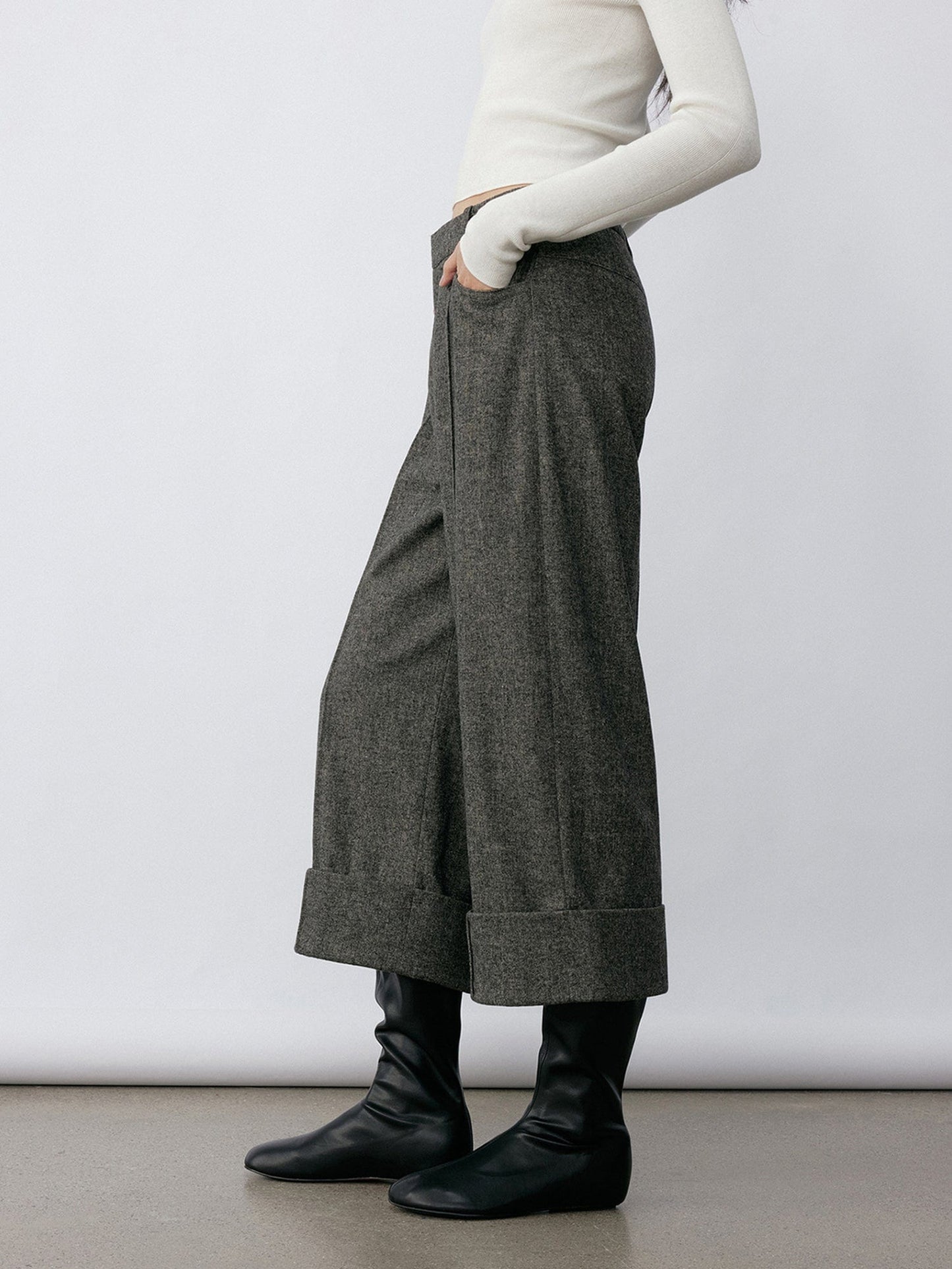 Woman wearing Arkin Studio turn-up hem wool trousers in gray and a white long sleeve top, with hands in pockets, wearing a pair of black shoes, standing sideways on a khaki floor, in front of a white wall