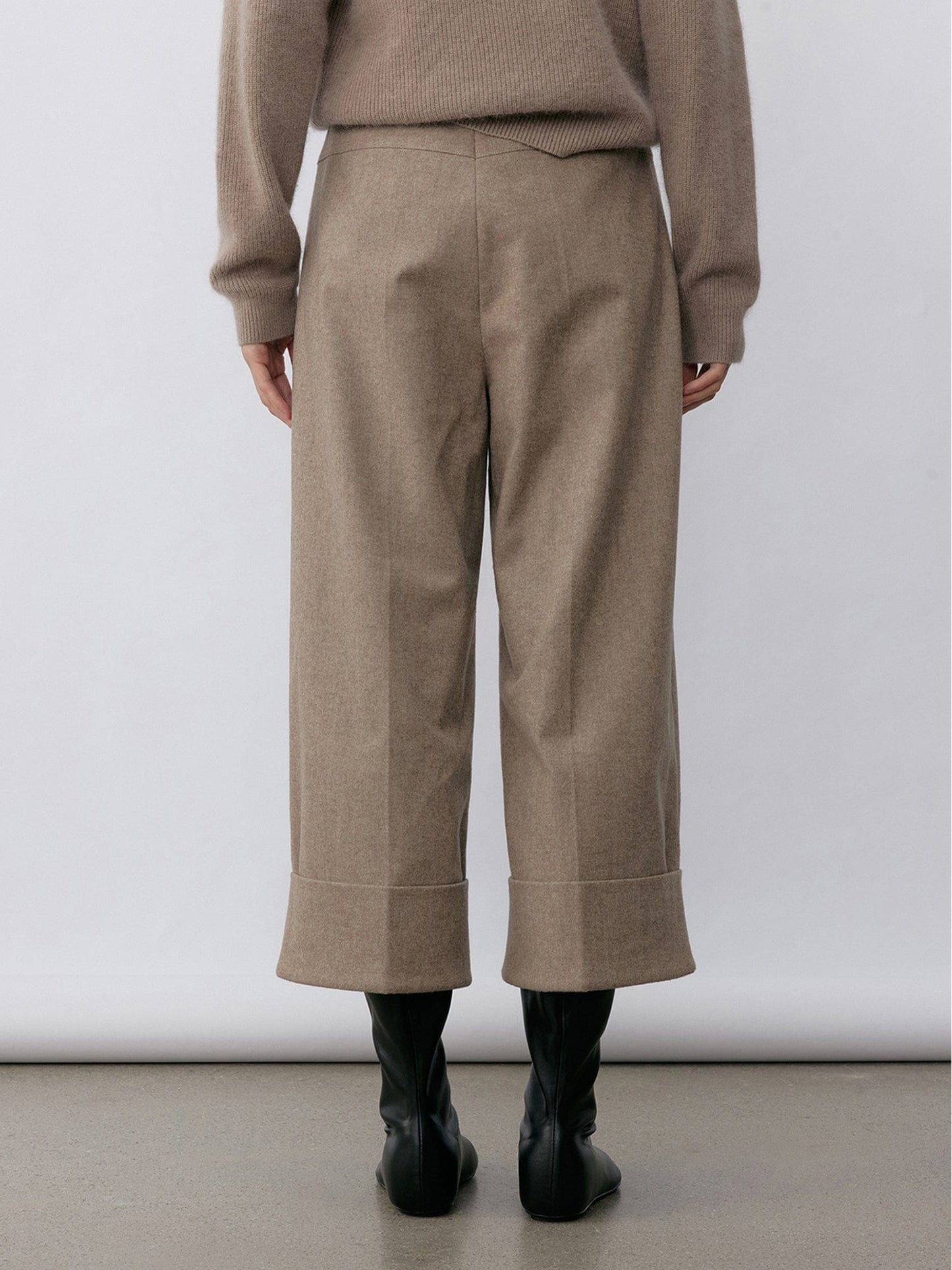 Woman wearing Arkin Studio turn-up hem wool trousers in taupe-gray with a black belt and matching knitwear, hands down at her sides, wearing a pair of black shoes, standing facing a white wall, on a white background