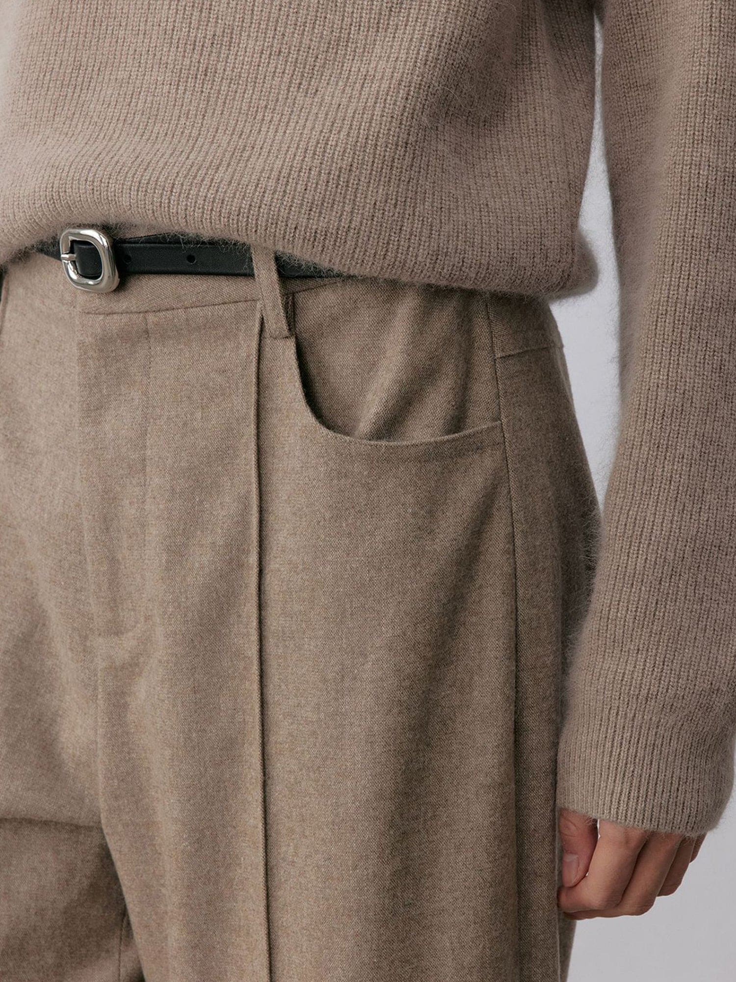 Close up of a woman wearing Arkin Studio turn-up hem wool trousers in taupe-gray with a black belt and matching knitwear, focus on her waist, standing facing the camera, on a white background