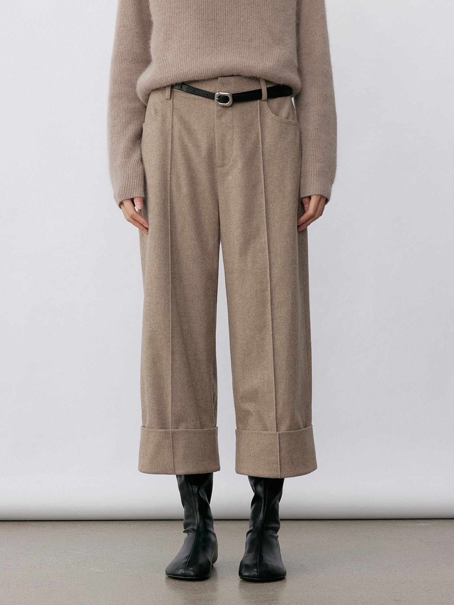Woman wearing Arkin Studio turn-up hem wool trousers in taupe-gray with a black belt and matching knitwear, hands down at her sides, wearing a pair of black shoes, standing facing the camera, on a white background