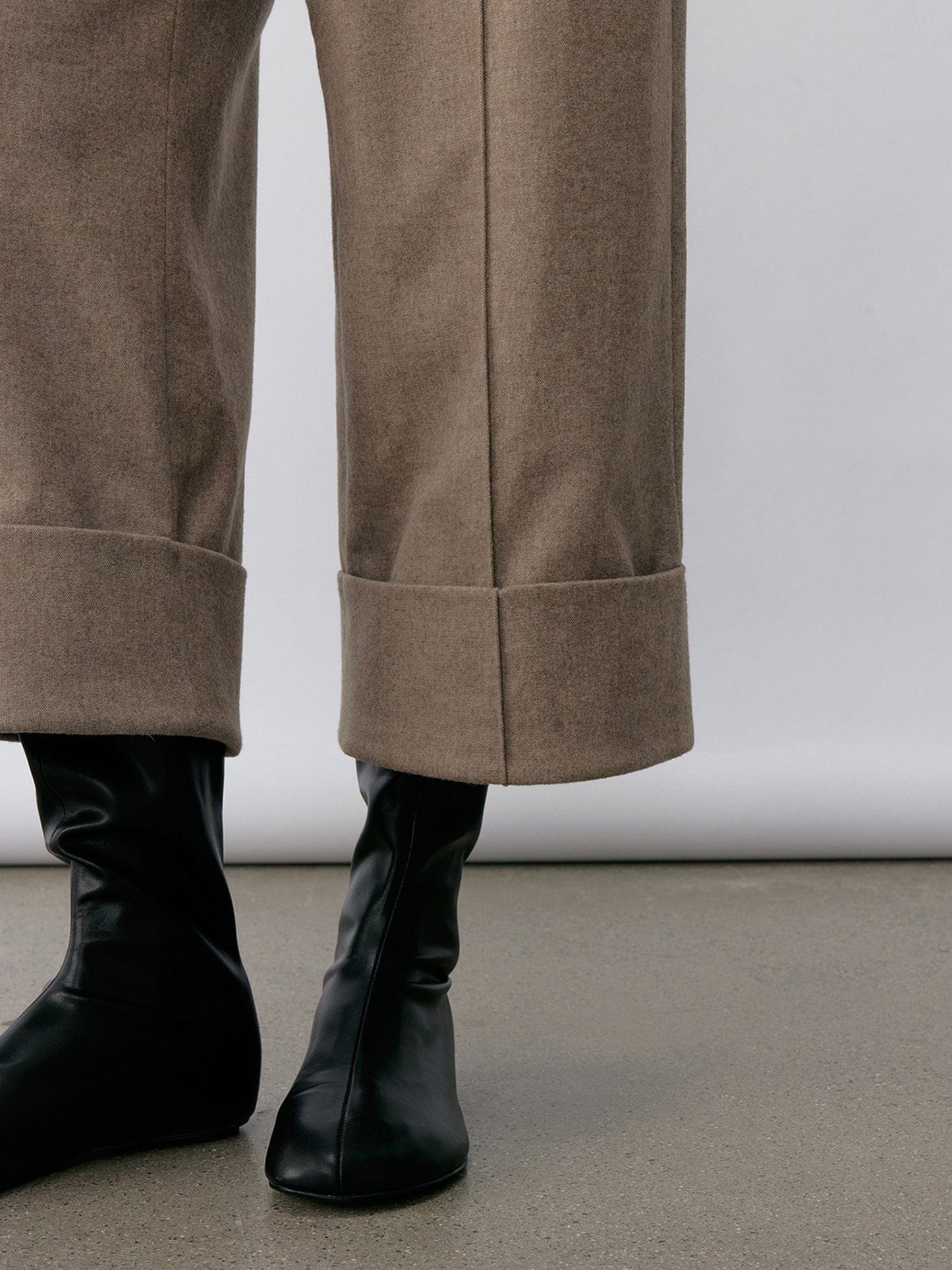 Close-up of a woman's legs from calf to feet wearing Arkin Studio turn-up hem wool trousers in taupe-gray with a pair of black shoes, standing facing the camera, on the khaki floor, in front of a white wall