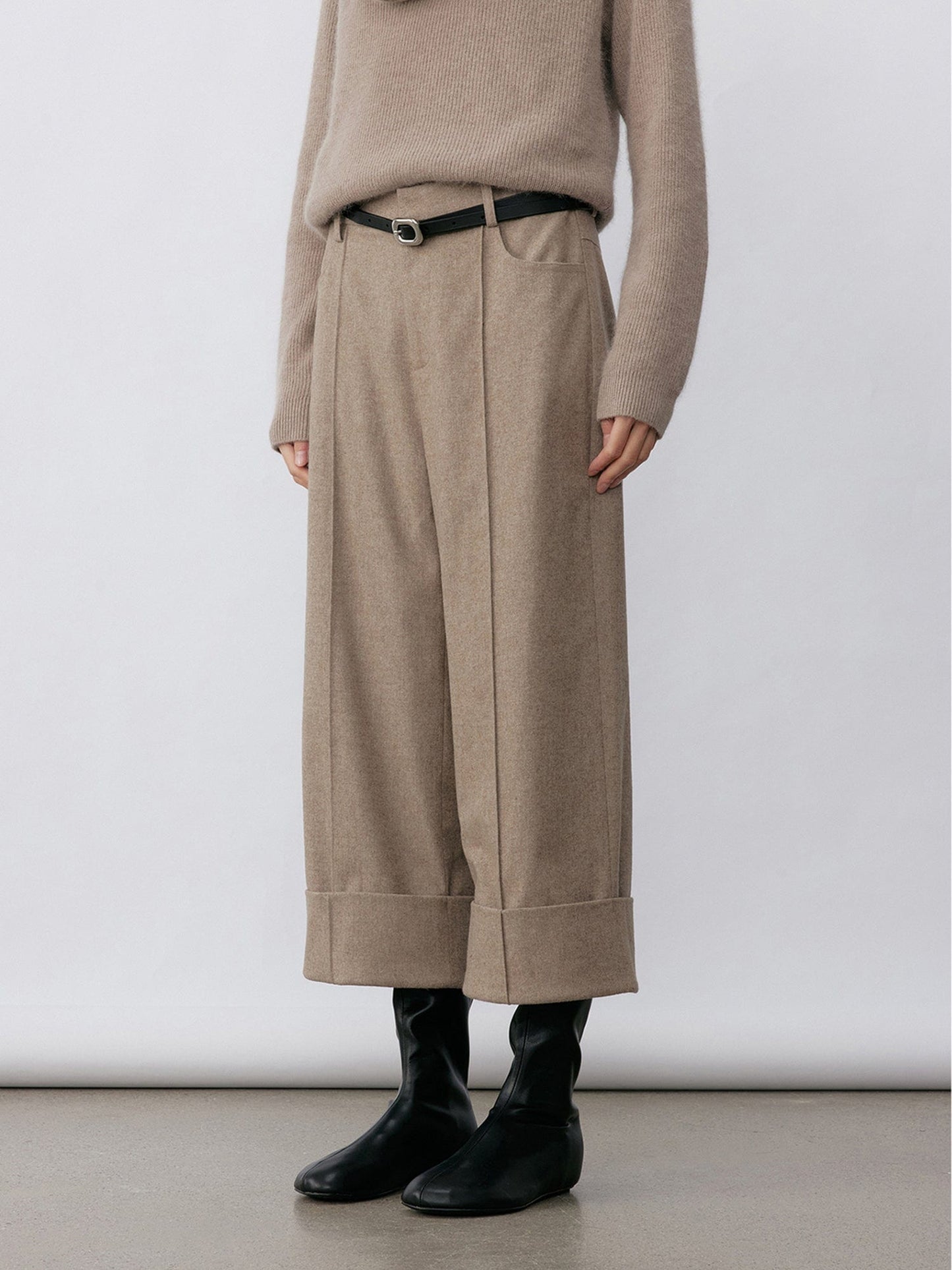 Woman wearing Arkin Studio turn-up hem wool trousers in taupe-gray with a black belt and matching knitwear, hands down at her sides, wearing a pair of black shoes, standing sideways, on a white background