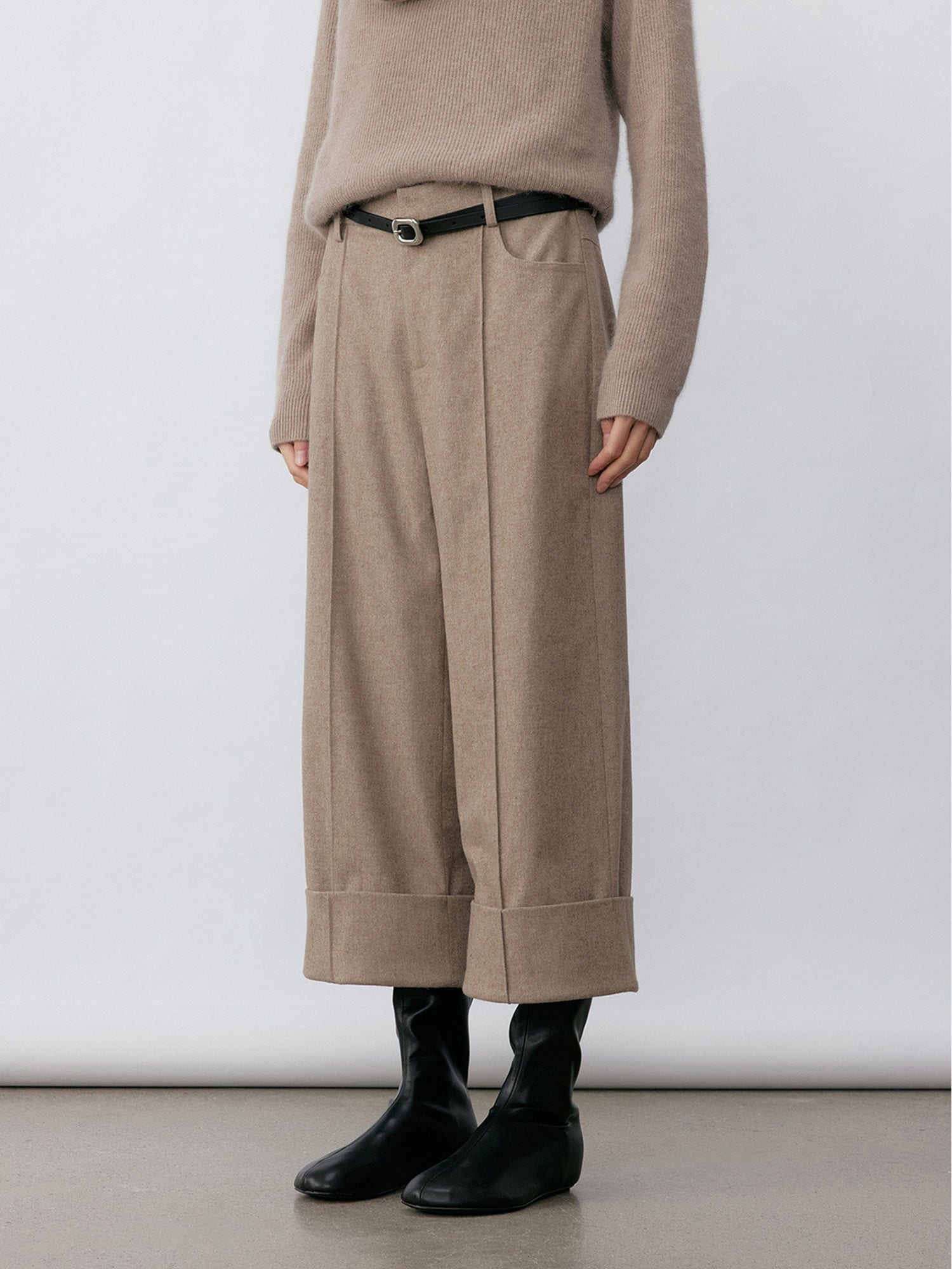 Woman wearing Arkin Studio turn-up hem wool trousers in taupe-gray with a black belt and matching knitwear, hands down at her sides, wearing a pair of black shoes, standing sideways, on a white background