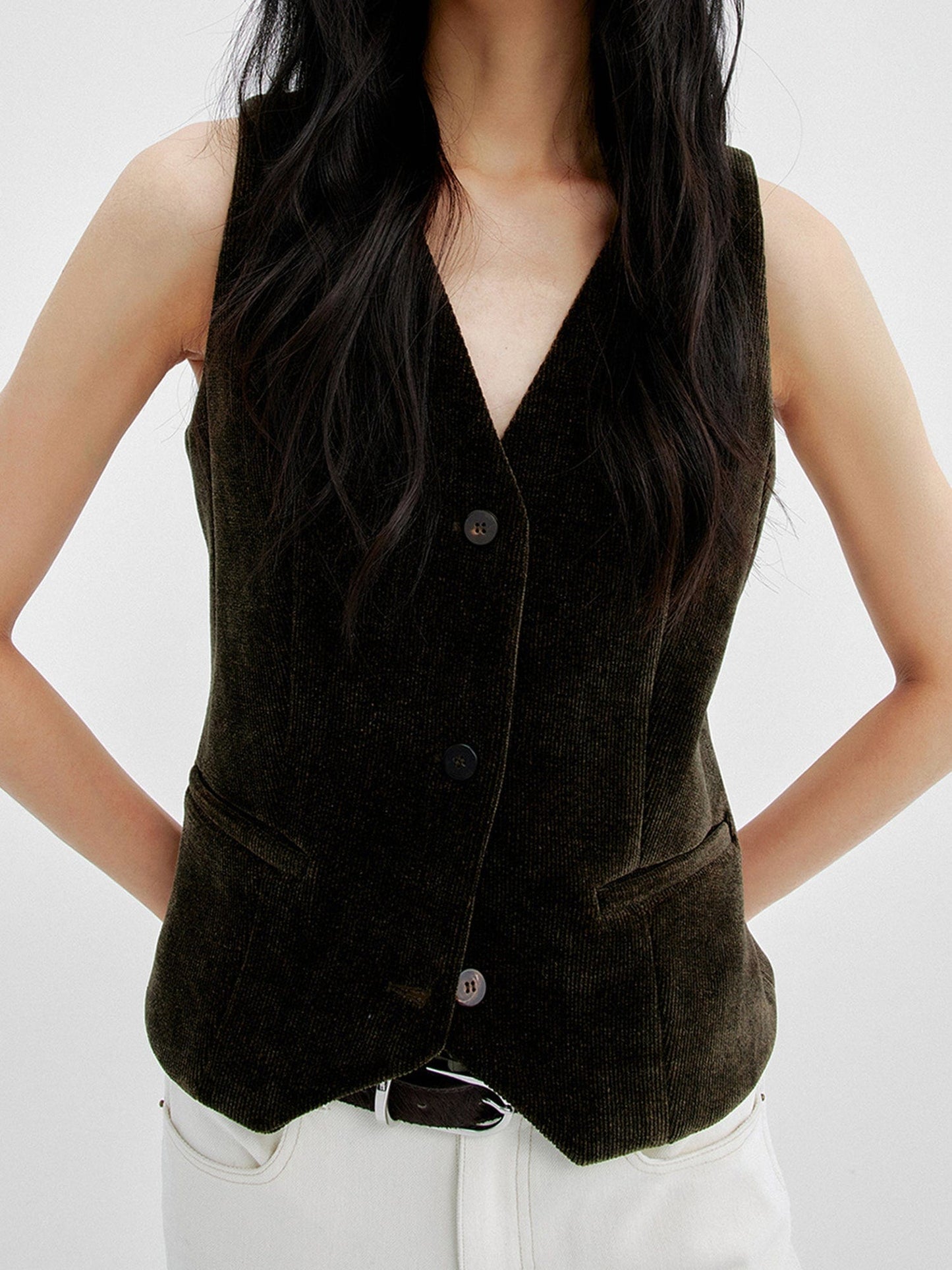 Woman wearing Arkin Studio v-neck corduroy vest in dark-olive and white pants, hands behind her back, standing facing the camera, on a white background