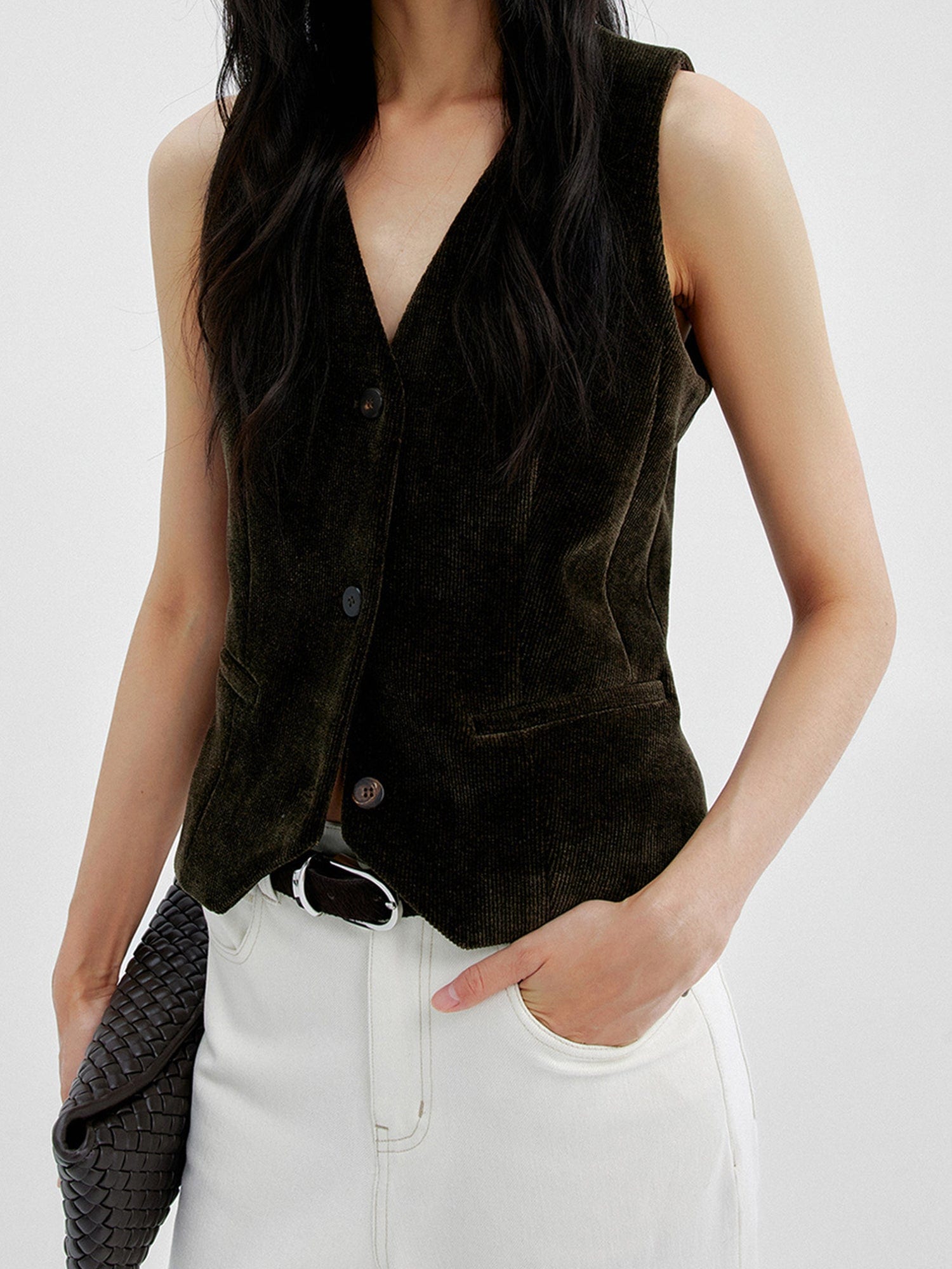 Woman wearing Arkin Studio v-neck corduroy vest in dark-olive and white pants, right hand holding a black bag and left hand in a pocket, standing facing the camera, on a white background