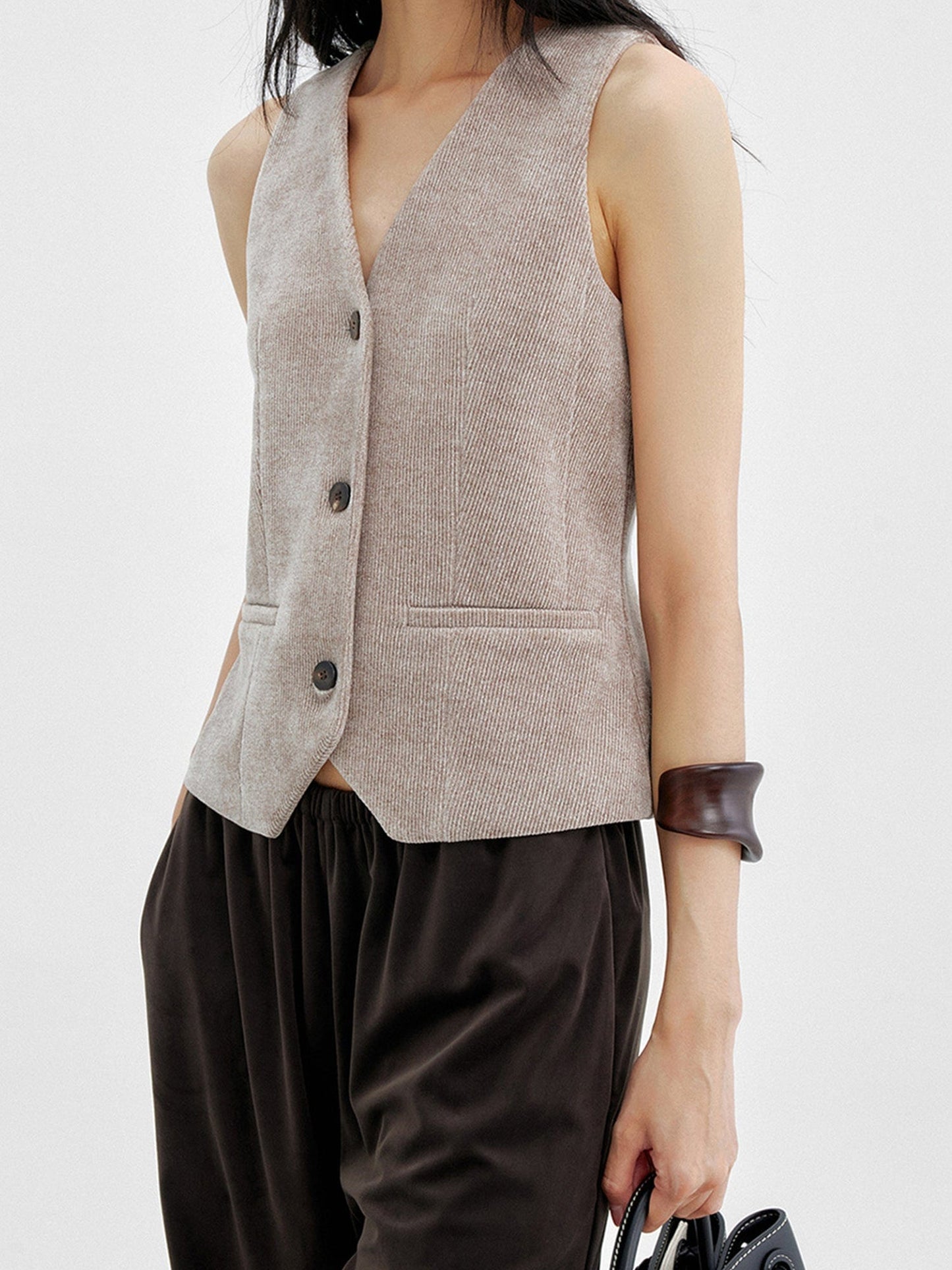 Woman wearing Arkin Studio v-neck corduroy vest in taupe and brown pants, holding a black bag, standing sideways, on a white background