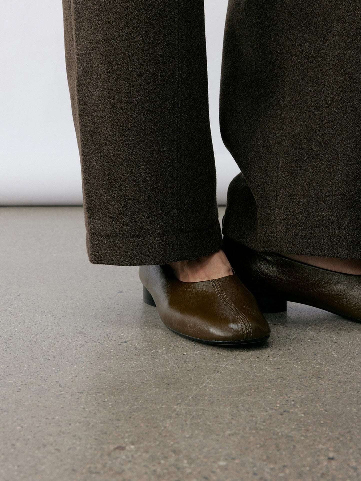 Close up of a woman wearing Arkin Studio wool blend straight leg trousers in olive-green and a pair of brown shoes, focus on her legs to shoes, standing sideways on the khaki floor, in front of a white wall