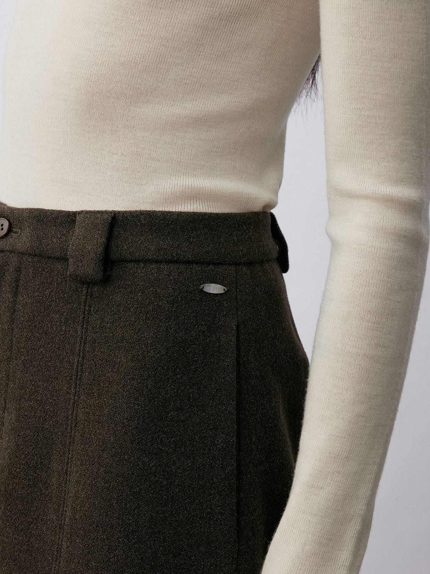 Close up of a woman wearing Arkin Studio wool blend straight leg trousers in olive-green and a white long sleeve top, focus on her waist, with one hand down at one side, standing facing the camera, on a white background
