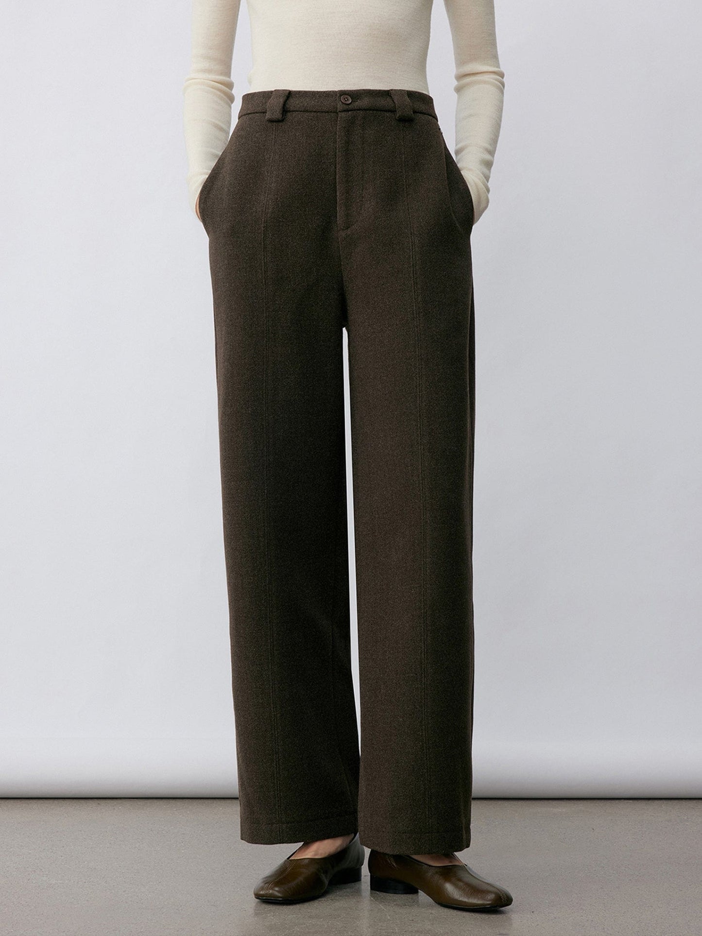 Woman wearing Arkin Studio wool blend straight leg trousers in olive-green and a white long sleeve top, wearing a pair of brown shoes, with hands in the pockets, standing facing the camera in front of a white wall, on a khaki floor
