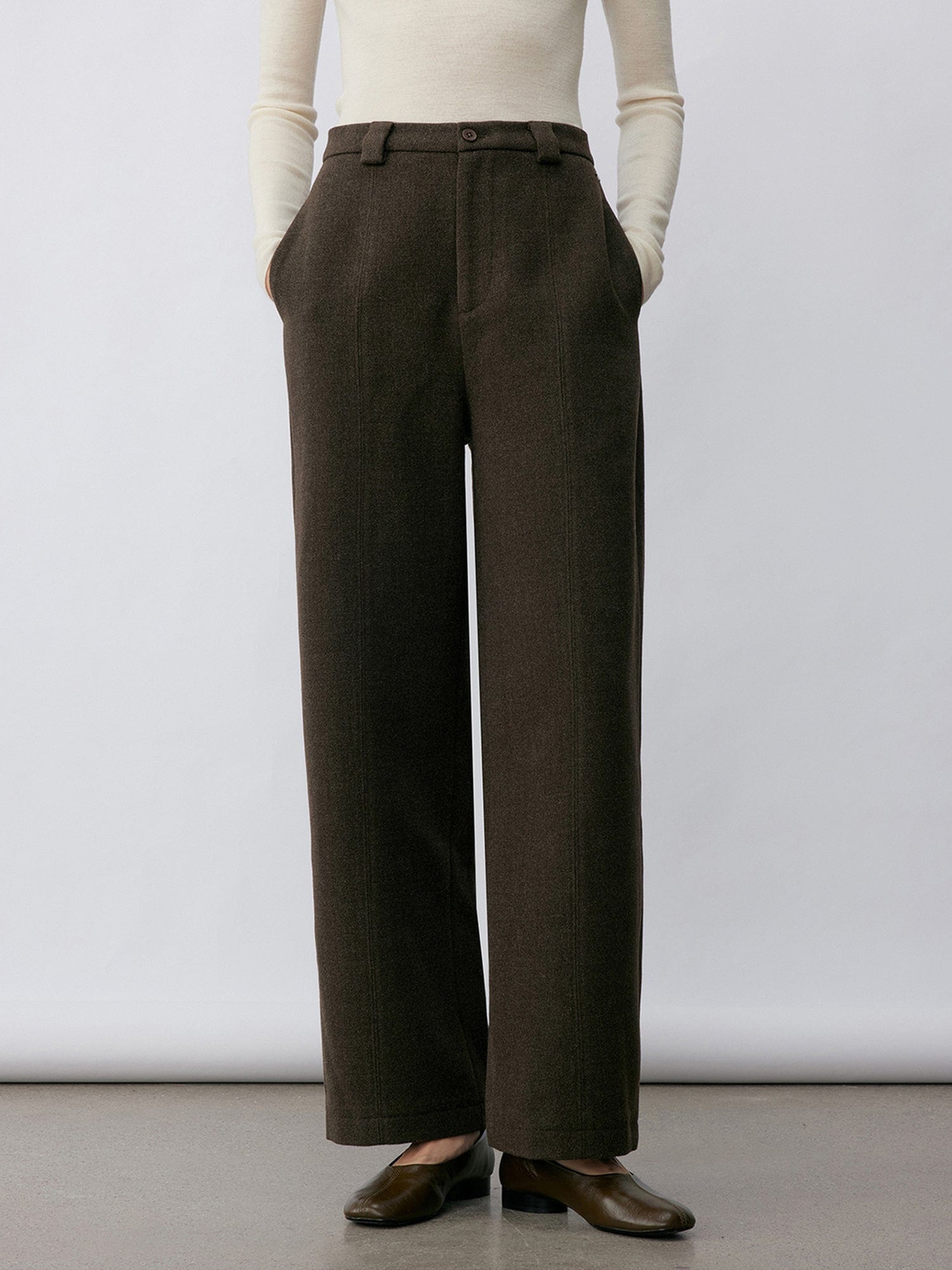 Woman wearing Arkin Studio wool blend straight leg trousers in olive-green and a white long sleeve top, wearing a pair of brown shoes, with hands in the pockets, standing facing the camera in front of a white wall, on a khaki floor