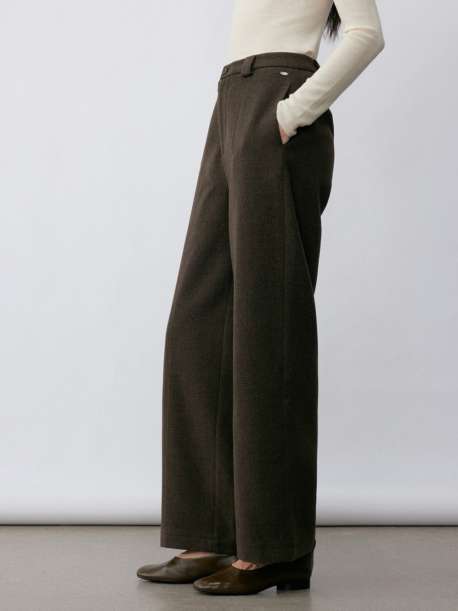 Woman wearing Arkin Studio wool blend straight leg trousers in olive-green and a white long sleeve top, wearing a pair of brown shoes, with hands in the pockets, standing sideways in front of a white wall, on a khaki floor