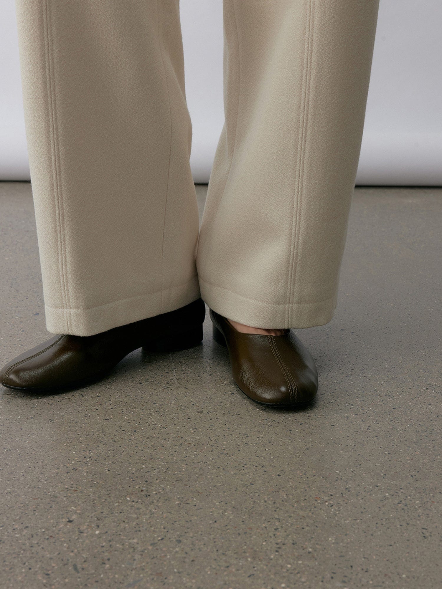 Close up of a woman wearing Arkin Studio wool blend straight leg trousers in white and a pair of brown shoes, focus on her legs to shoes, standing facing the camera on the khaki floor, in front of a white wall