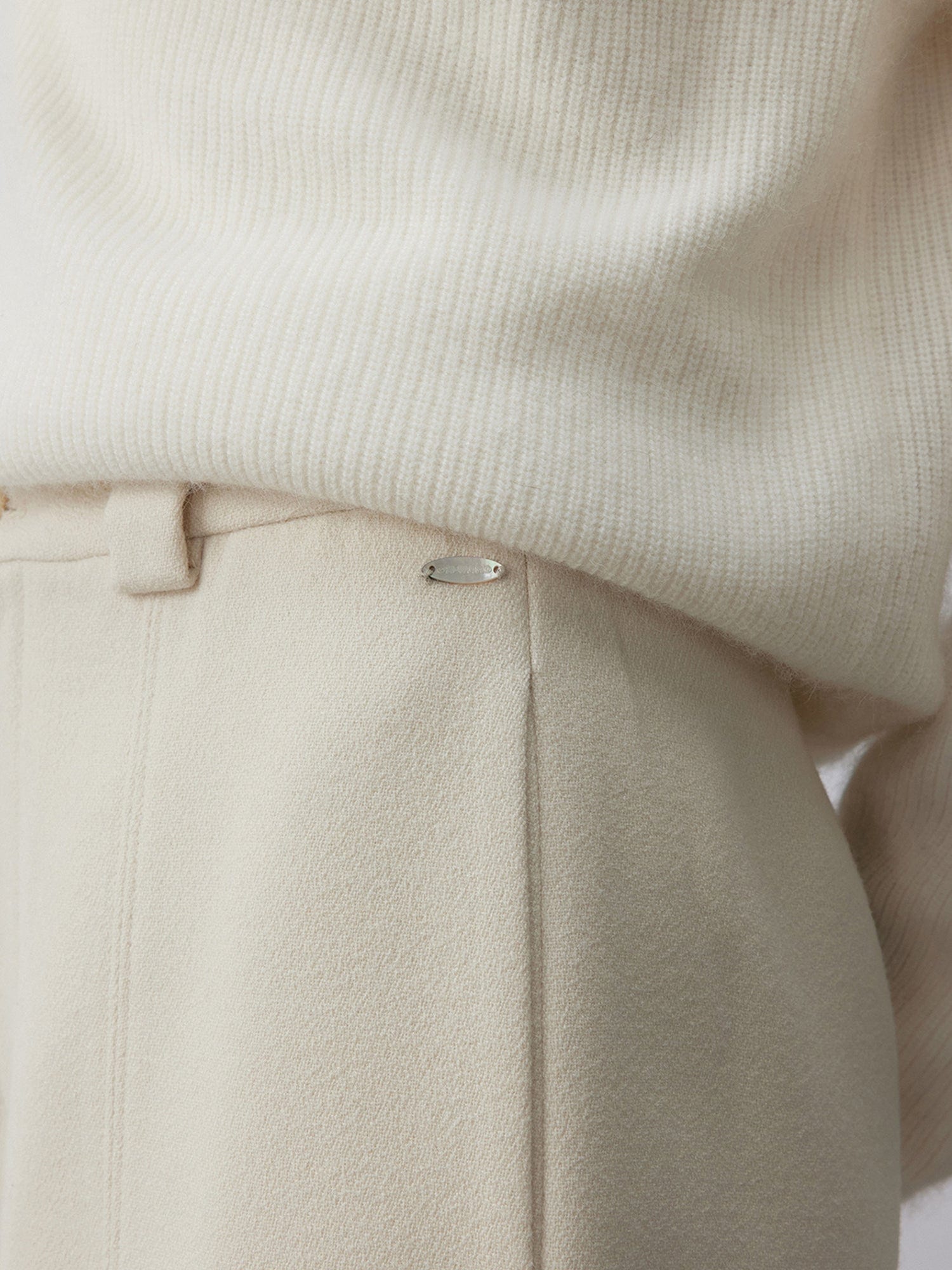 Close up of a woman wearing Arkin Studio wool blend straight leg trousers in white and a white knitwear, focus on her waist, with one hand behind her back, standing facing the camera