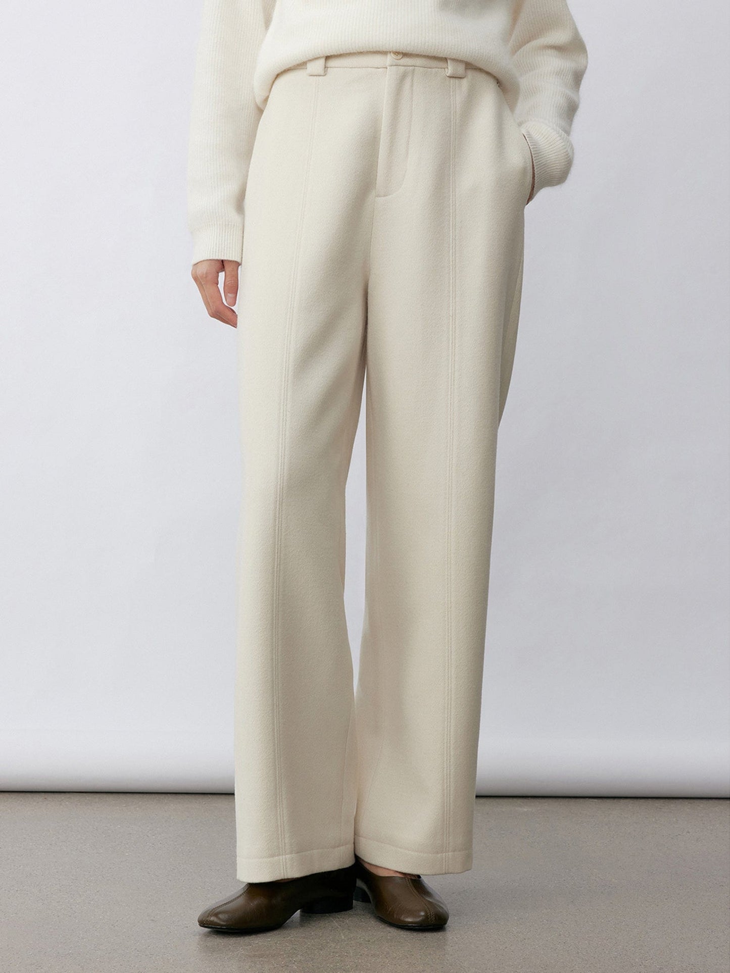 Woman wearing Arkin Studio wool blend straight leg trousers in white and a white long sleeve knitwear, wearing a pair of brown shoes, with one hand in a pocket, standing facing the camera in front of a white wall, on a khaki floor