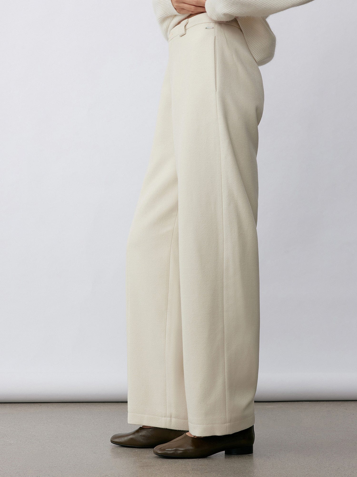 Woman wearing Arkin Studio wool blend straight leg trousers in white and a white long sleeve knitwear, wearing a pair of brown shoes, standing sideways in front of a white wall, on a khaki floor